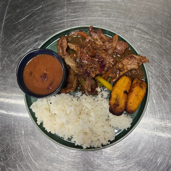 A plate of steak with onions and peppers, white rice, fried plantains, and a side of beans.