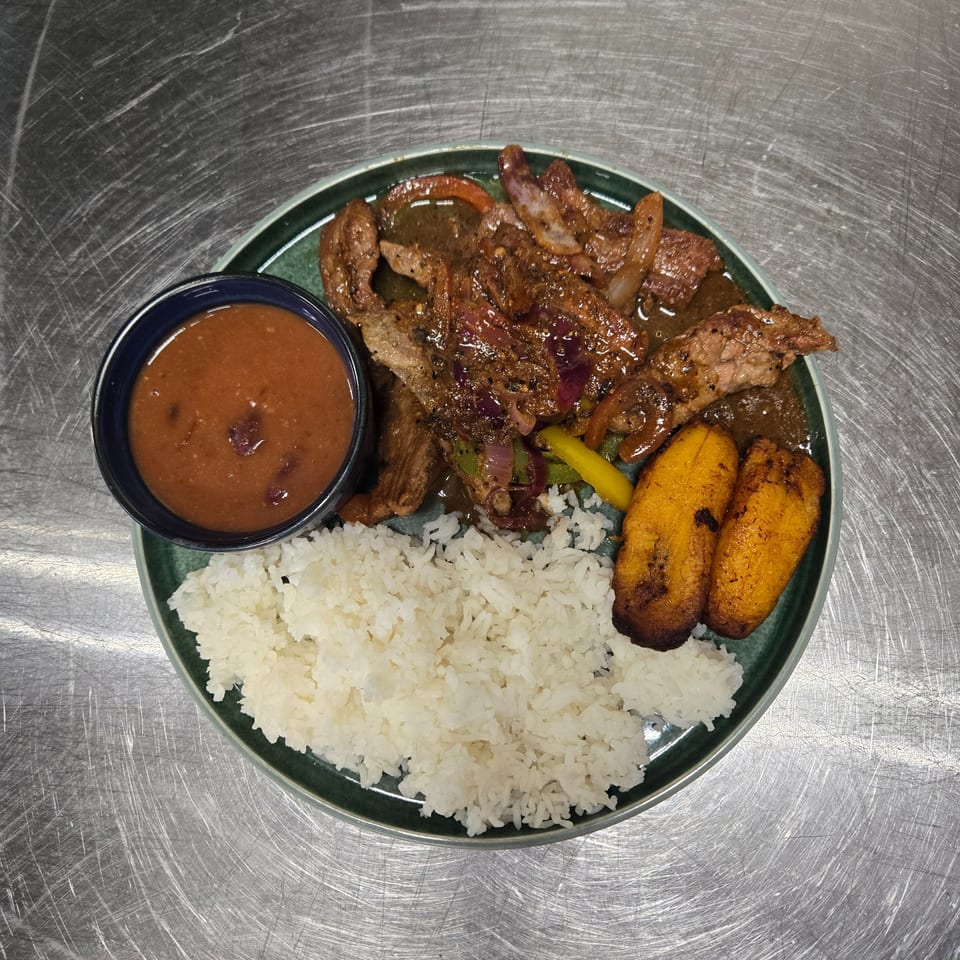 A plate of steak with onions and peppers, white rice, fried plantains, and a side of beans.
