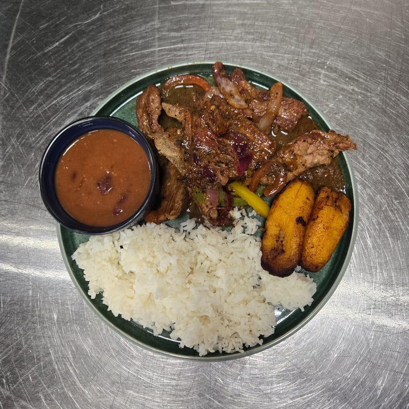 A plate of steak with onions and peppers, white rice, fried plantains, and a side of beans.