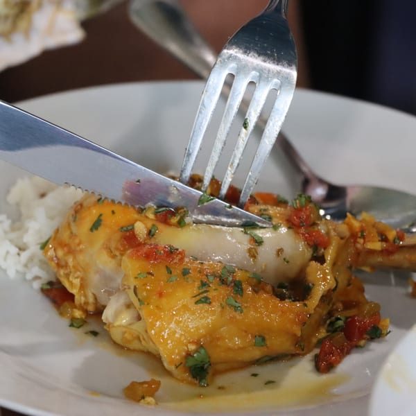 A knife and fork cutting into a piece of chicken with sauce and rice on a white plate.