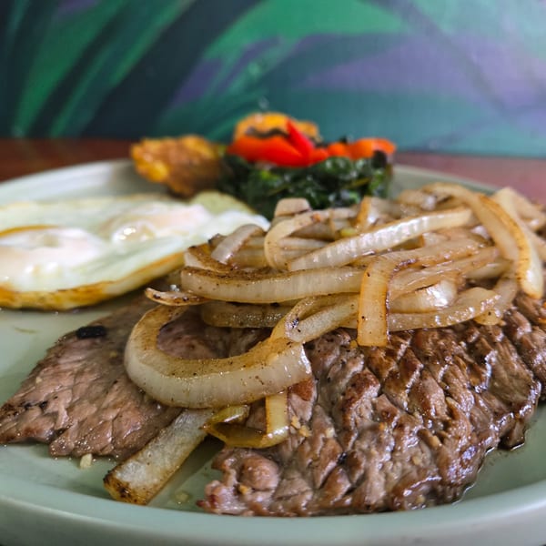 Steak with caramelized onions, two fried eggs, plantains, and greens with red peppers.