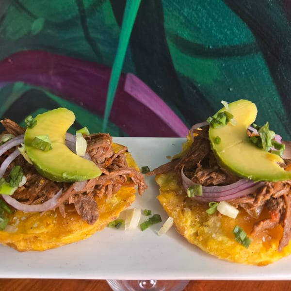Two plantain tostones topped with shredded beef, red onion, avocado slices, and green onions.