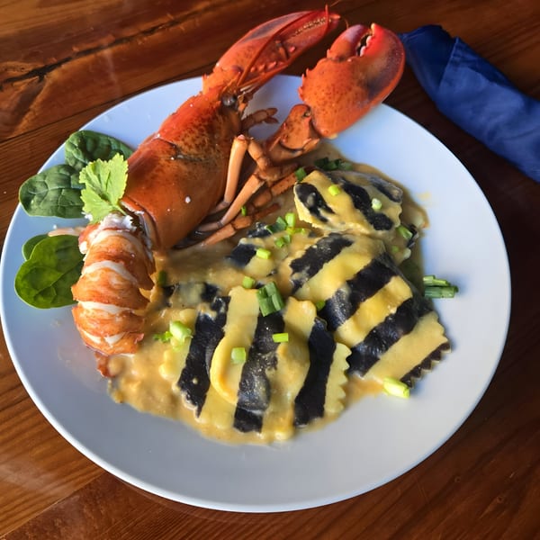 A whole cooked lobster and striped ravioli in sauce, garnished with greens on a white plate.