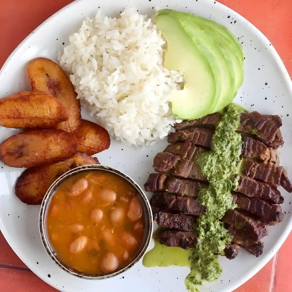 Latin-American dish with sliced steak, green sauce, rice, avocado, fried plantains, and beans.