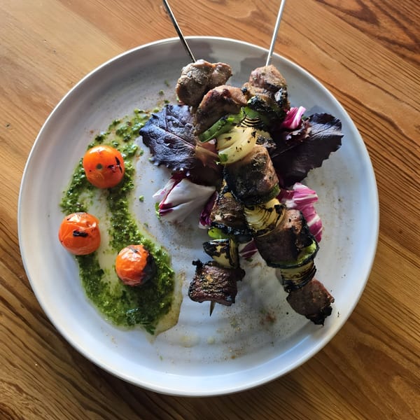 Two skewers of grilled meat and vegetables with roasted tomatoes and green sauce on a white plate.